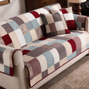 May include: A patchwork sofa cover and two matching throw pillows. The cover features a quilted design with squares of red, blue, beige, and brown. The pillows echo the same pattern. The sofa has a light beige frame.