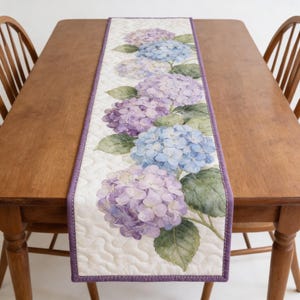 Puede incluir: Camino de mesa acolchado con un diseño floral de hortensias en tonos morados y azules. El camino de mesa tiene un fondo blanco con un patrón ondulado y un borde morado. El camino de mesa se muestra sobre una mesa de madera.