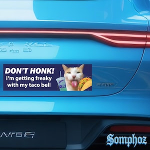 May include: A blue car bumper sticker with the text "DON'T HONK! I'm getting freaky with my taco bell." The sticker features a cat with a taco and a Taco Bell drink. The word "Somphoz" is written in a stylized font.
