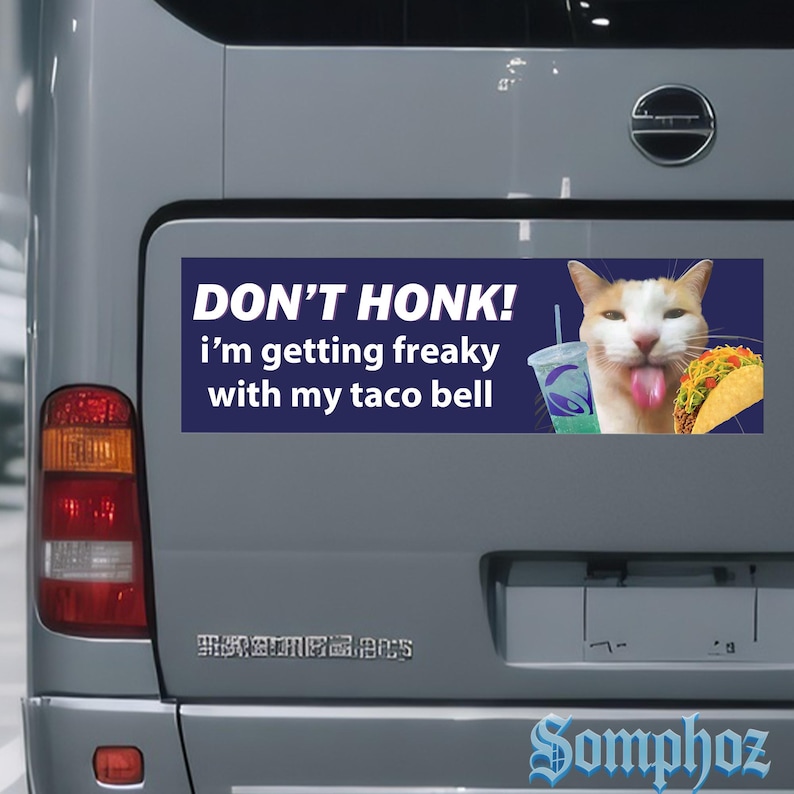 May include: A rectangular bumper sticker with the text "DON'T HONK! I'm getting freaky with my taco bell." A cartoon cat with its tongue out is next to a Taco Bell drink and taco. The sticker is on the back of a vehicle.