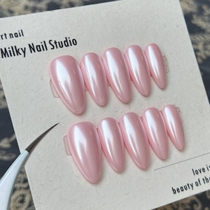 May include: A set of ten almond-shaped press-on nails with a glossy, pearlescent light pink finish. The nails are displayed on a cream-colored card labeled Milky Nail Studio. A white-handled metal tool is positioned on the left side of the card.