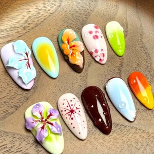 May include: Ten artificial nails with diverse designs are displayed. The nails showcase a variety of colours, including blue, yellow, orange, green, and pink. Some nails feature floral decorations, while others have abstract patterns and water droplet details. The nails are presented on a wooden surface.