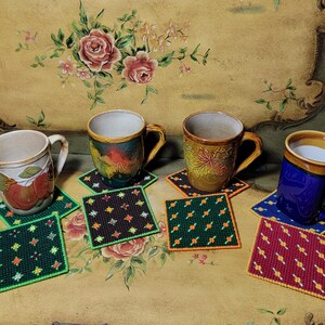 May include: Four ceramic mugs in various colors, each resting on a square, needlepoint coaster. The mugs are blue, brown, and cream-colored, with floral and abstract designs. The coasters are green, black, and red with star patterns.