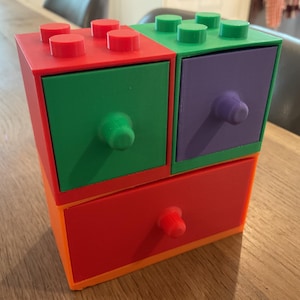 Könnte beinhalten: Ein vier Schubladen Organizer aus Kunststoff mit einem roten, grünen, violetten und orangen Farbschema. Die Schubladen haben ein Lego-ähnliches Design mit runden Knöpfen.