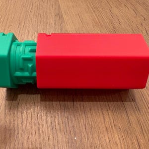 Op de afbeelding: Een groen en rood rechthoekig object. Het groene uiteinde heeft een geometrisch ontwerp. Het rode gedeelte is een effen, gladde rechthoek. Het object ligt op een houten oppervlak.