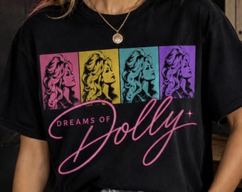 May include: A black short-sleeved t-shirt featuring a graphic print of four stylized portraits of a woman with blonde hair in pink, yellow, teal, and purple squares. Below the portraits, the text reads "DREAMS OF Dolly" in pink script.