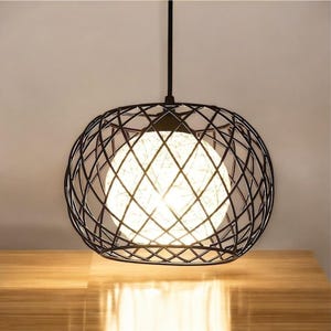 Peut inclure: Un luminaire suspendu en métal noir avec un abat-jour rond en cage de fil. La lumière est allumée et éclaire la pièce d'une lueur blanche chaude.