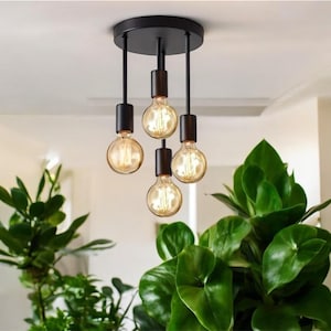 Könnte beinhalten: Eine schwarze Deckenleuchte mit vier hängenden Glühbirnen. Die Glühbirnen sind aus klarem Glas mit einem Filament-Design und strahlen ein warmes weißes Licht ab. Die Leuchte ist an einer weißen Decke montiert.
