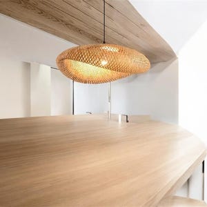 May include: A woven bamboo pendant light fixture hangs from a ceiling in a modern interior design setting. The light fixture is oval shaped and has a natural wood colour.