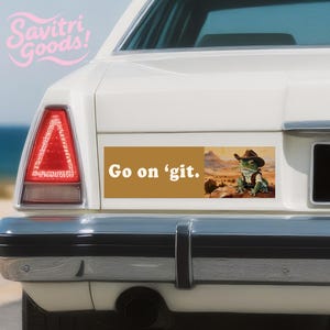 May include: A rectangular bumper sticker with the text "Go on 'git." features a frog wearing a cowboy hat and vest, set against a desert landscape. The sticker is affixed to the back of a white car.