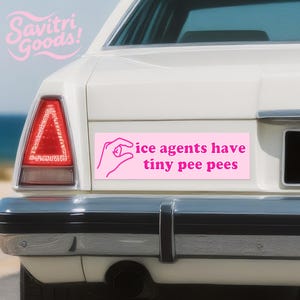 May include: A pink bumper sticker on a white car with the text "ice agents have tiny pee pees" in a pink font. The sticker also features a pink graphic of a hand pinching a small object. The car has a red tail light.