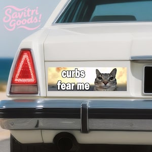 Könnte beinhalten: Heckseite eines weißen Autos mit einem Aufkleber mit der Aufschrift "curbs fear me" über dem Gesicht einer Katze. Der Aufkleber hat einen Sonnenuntergangshintergrund. Das Logo Savitri Goods! befindet sich in der oberen linken Ecke.