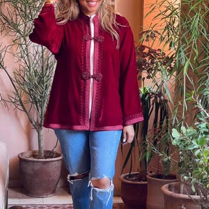 Burgundy red Moroccan velvet jacket featuring hand-embroidered trim and traditional frogging closures, worn with ripped blue jeans and sunglasses in bohemian courtyard setting with tropical greenery and terracotta planters