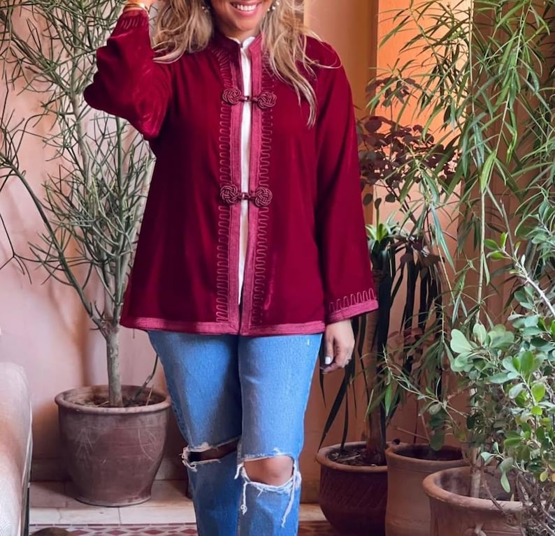 Burgundy red Moroccan velvet jacket featuring hand-embroidered trim and traditional frogging closures, worn with ripped blue jeans and sunglasses in bohemian courtyard setting with tropical greenery and terracotta planters