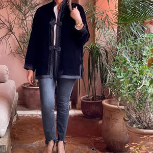 Black handcrafted Moroccan velvet jacket with contrasting embroidered detailing and ornate knotted buttons, styled casually with blue denim jeans and black pumps in Marrakech terrace surrounded by potted plants and traditional Moroccan architecture