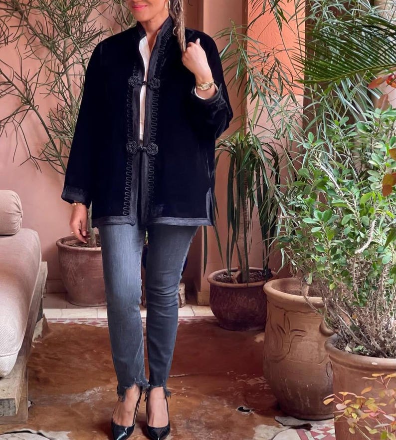 Black handcrafted Moroccan velvet jacket with contrasting embroidered detailing and ornate knotted buttons, styled casually with blue denim jeans and black pumps in Marrakech terrace surrounded by potted plants and traditional Moroccan architecture