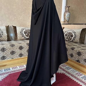 May include: A long, black hooded cloak. The cloak is made of a flowing fabric and drapes to the floor. The hood is up, and the cloak is worn over white clothing. The background includes a red rug and patterned pillows.