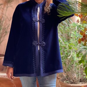 Handmade Moroccan navy blue velvet jacket with intricate embroidered detailing, traditional knotted closures, and decorative passementerie trim along front opening, styled with light blue jeans and palm frond accessory in sunny Marrakech courtyard with terracotta walls and potted plants