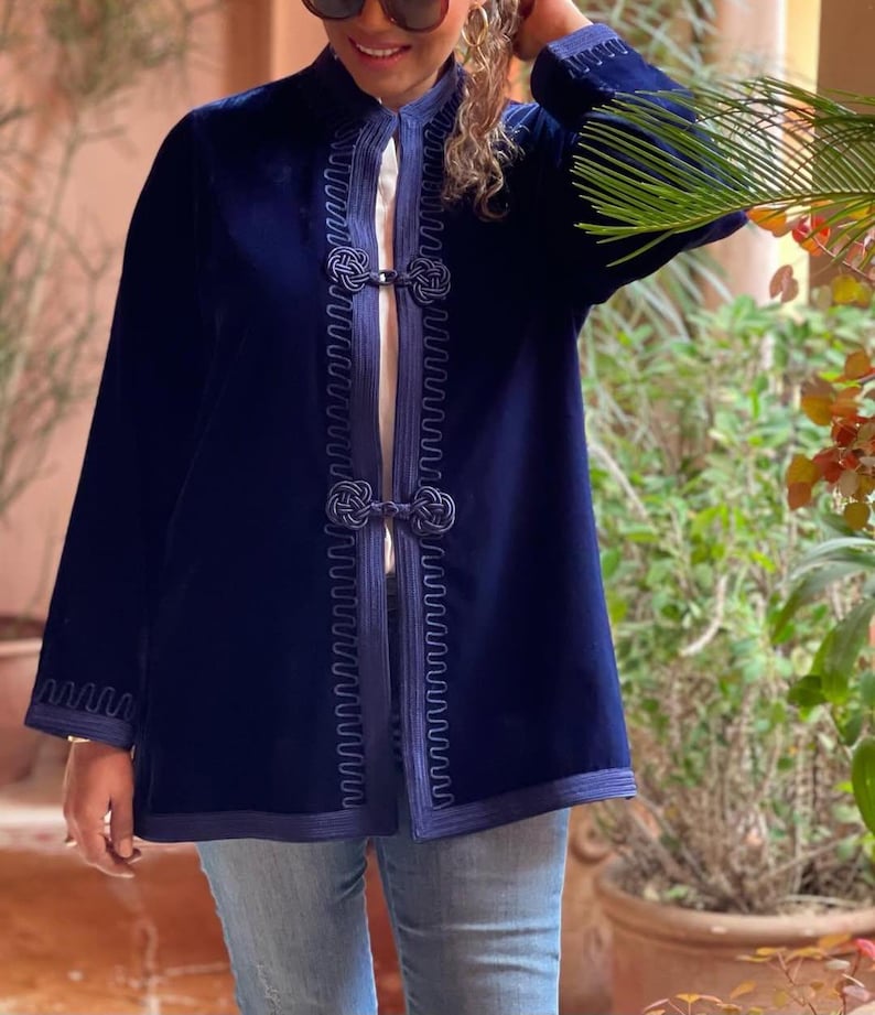 Handmade Moroccan navy blue velvet jacket with intricate embroidered detailing, traditional knotted closures, and decorative passementerie trim along front opening, styled with light blue jeans and palm frond accessory in sunny Marrakech courtyard with terracotta walls and potted plants