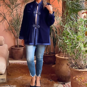 Full body view of woman wearing traditional Moroccan navy blue velvet caftan jacket with embroidered geometric patterns, paired with distressed jeans and black platform heels, standing in authentic Moroccan riad with desert plants and orange adobe walls