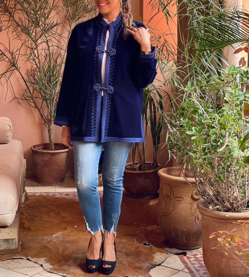 Full body view of woman wearing traditional Moroccan navy blue velvet caftan jacket with embroidered geometric patterns, paired with distressed jeans and black platform heels, standing in authentic Moroccan riad with desert plants and orange adobe walls