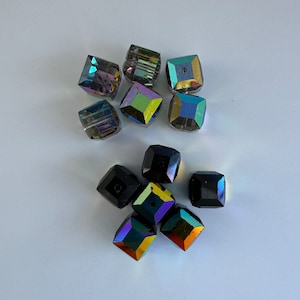May include: Assortment of iridescent glass beads in square shapes. Some beads are black, while others are a mix of green, blue, and purple.