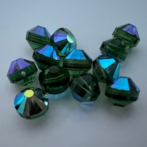 May include: A group of dark green faceted glass beads with iridescent blue and purple accents. The beads are scattered on a white surface.