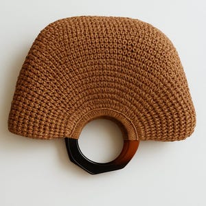 May include: A brown, fan-shaped handbag with a woven texture. The bag features a circular handle made of dark brown and black geometric-shaped plastic. The bag is set against a white background.