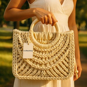 May include: A cream-colored woven tote bag with a wooden handle. The bag has a unique design with a braided pattern and a small tag that says "ecrafworkshop".