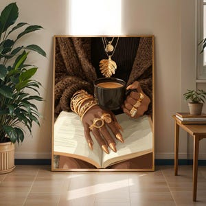 May include: A framed wall art print depicting a person wearing a dark brown textured sweater, multiple gold bracelets, and gold rings, holding a black mug filled with coffee over an open book. A gold leaf pendant necklace hangs around the neck.