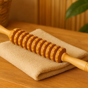 Puede incluir: Un rodillo de masaje de madera con ruedas estriadas, apoyado sobre una toalla beige. El rodillo es de color marrón claro y está diseñado para la terapia muscular. El fondo incluye una superficie de madera y una cesta de mimbre.
