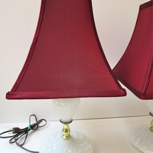 May include: Pair of vintage table lamps with burgundy shades. The lamps feature a white, textured base and a gold-colored stem. The shades are a deep burgundy color, and the lamps are plugged in with a brown cord.
