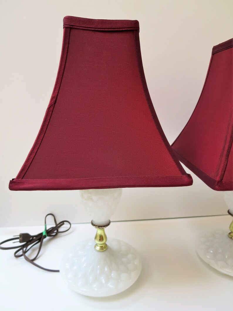 May include: Pair of vintage table lamps with burgundy shades. The lamps feature a white, textured base and a gold-colored stem. The shades are a deep burgundy color, and the lamps are plugged in with a brown cord.