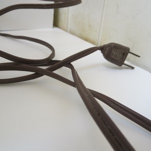 May include: A brown electrical cord with a matching plug. The cord is coiled and lies on a white surface. The plug has two prongs and is made of a dark brown plastic.
