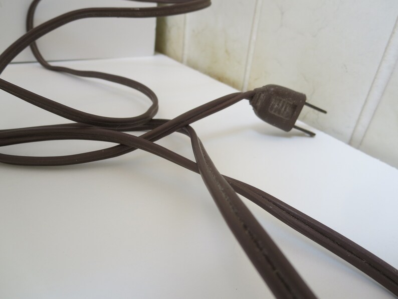 May include: A brown electrical cord with a matching plug. The cord is coiled and lies on a white surface. The plug has two prongs and is made of a dark brown plastic.
