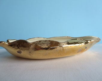 May include: A gold-colored, oblong ceramic bowl with a scalloped rim. The interior is white, and the exterior has a metallic gold finish with some areas of wear. The bowl is set against a light blue background.
