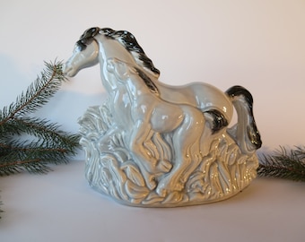 May include: A ceramic sculpture of two horses in a running pose, with a light gray glaze and black manes and tails. The horses are set on a base with a textured, grassy design. Decorative item.