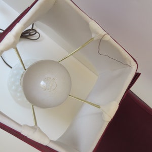 May include: A burgundy lampshade with a square shape and white interior. The lampshade is supported by brass-colored rods and surrounds a white, frosted glass lightbulb. A brown electrical cord is visible.
