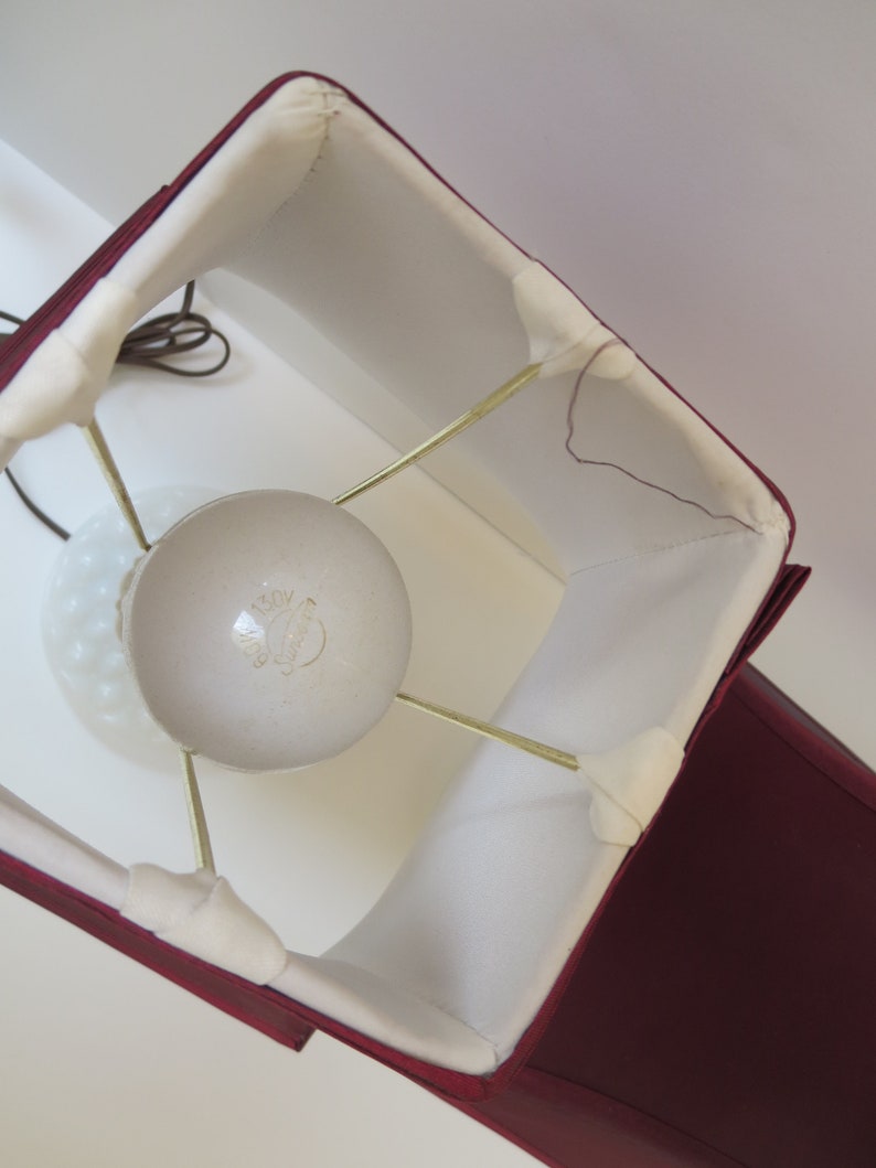 May include: A burgundy lampshade with a square shape and white interior. The lampshade is supported by brass-colored rods and surrounds a white, frosted glass lightbulb. A brown electrical cord is visible.