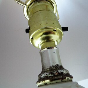 May include: Close-up of a lamp fixture, featuring a gold-colored bulb socket and a white ceramic base. The socket has a black on/off switch. The base shows signs of age with some discoloration.