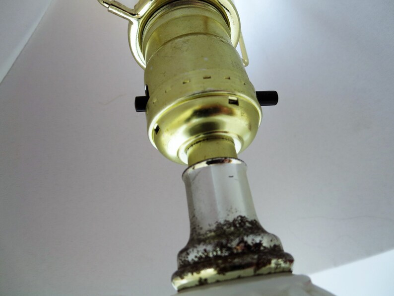 May include: Close-up of a lamp fixture, featuring a gold-colored bulb socket and a white ceramic base. The socket has a black on/off switch. The base shows signs of age with some discoloration.