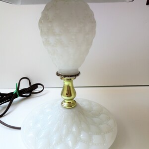 May include: A white, vintage-style table lamp with a textured, bulbous body and a round base. The lamp features a gold-colored metal stem and a white lampshade. The power cord is brown and has a green twist tie.