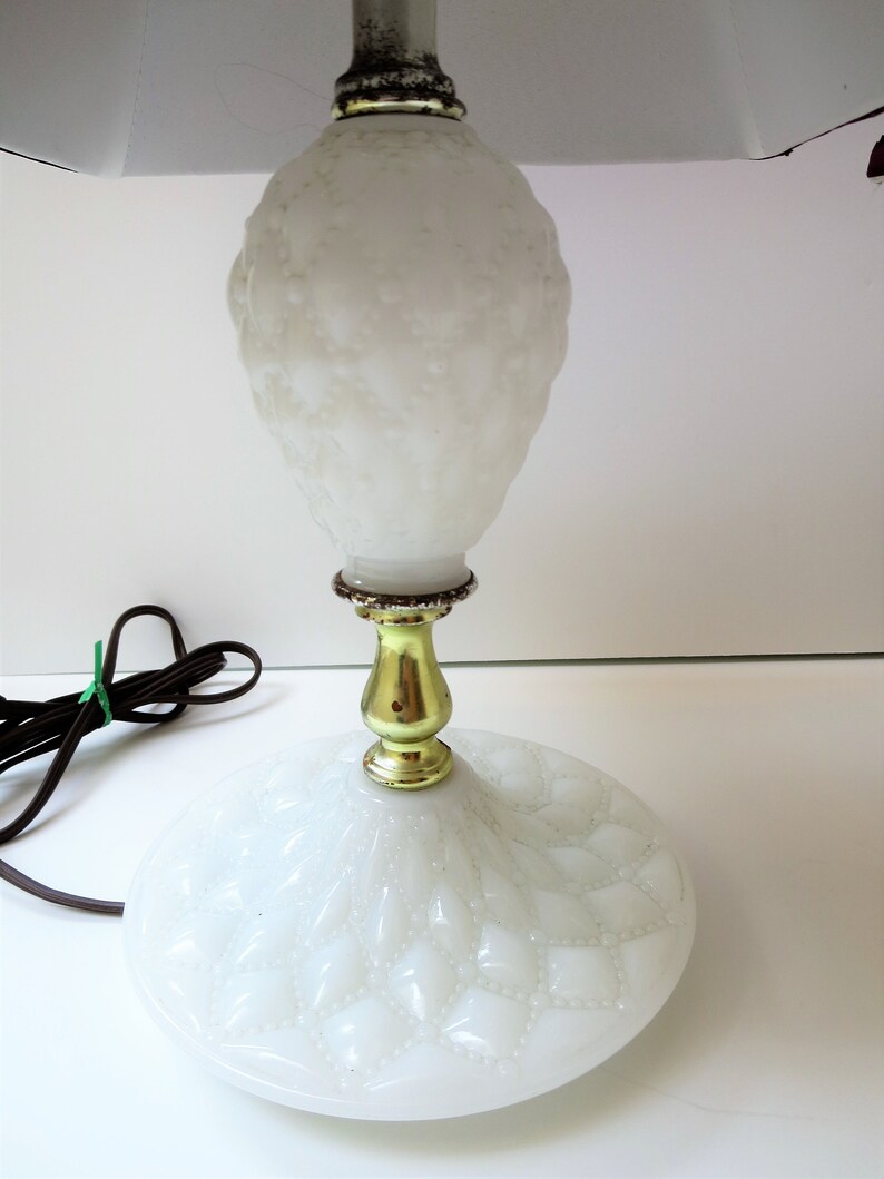 May include: A white, vintage-style table lamp with a textured, bulbous body and a round base. The lamp features a gold-colored metal stem and a white lampshade. The power cord is brown and has a green twist tie.