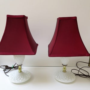 May include: Two matching table lamps with burgundy shades. The lamps have white, textured bases and gold-colored accents. The shades are square-shaped and the lamps are plugged in.