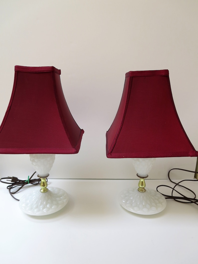 May include: Two matching table lamps with burgundy shades. The lamps have white, textured bases and gold-colored accents. The shades are square-shaped and the lamps are plugged in.