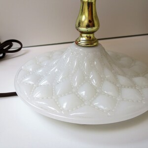 May include: Close-up of a white glass lamp base with a diamond pattern and a gold-colored metal stem. The base has a round shape and a textured surface. A brown electrical cord is visible.