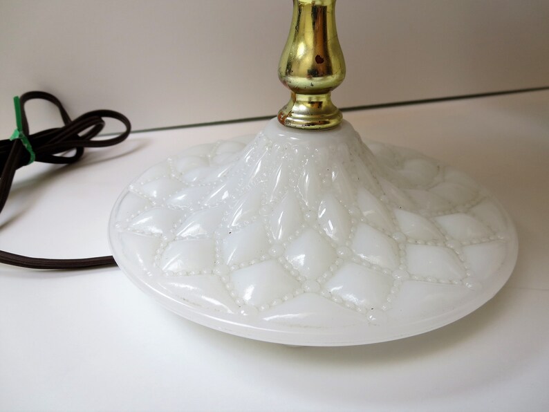May include: Close-up of a white glass lamp base with a diamond pattern and a gold-colored metal stem. The base has a round shape and a textured surface. A brown electrical cord is visible.