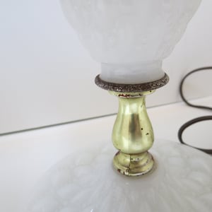 May include: A vintage white glass lamp with a textured shade and base. The lamp features a gold-colored metal stem and a decorative ring. A brown electrical cord is visible.