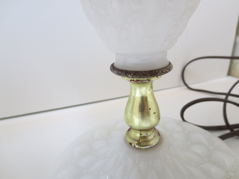 May include: A vintage white glass lamp with a textured shade and base. The lamp features a gold-colored metal stem and a decorative ring. A brown electrical cord is visible.