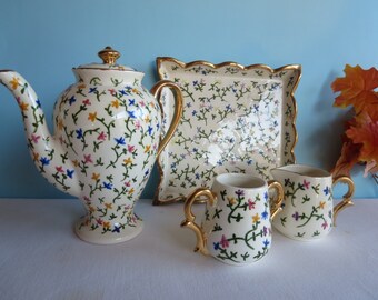 May include: A vintage porcelain tea set featuring a teapot, serving tray, creamer, and sugar bowl. The set is decorated with a delicate floral pattern in blue, pink, and yellow on a cream background. Gold trim accents the edges and handles.